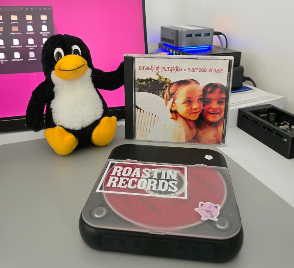 Estojo do CD Siamese Dream na mesa, ao lado de um pinguim Tux de pelúcia e de um CD player portátil com o disco vermelho aparecendo pela tampa